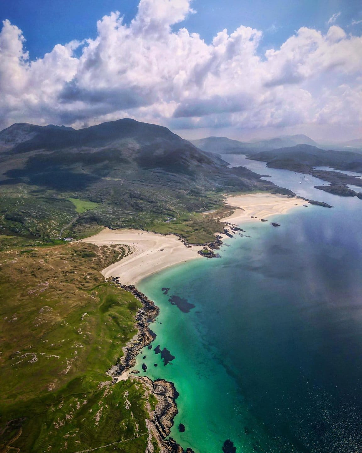 Aerial view of Silver Strand Beach in Ireland, showcasing a sweeping sandy coastline bordered by clear turquoise waters, rugged rocky shores, and rolling green hills under a partly cloudy sky.