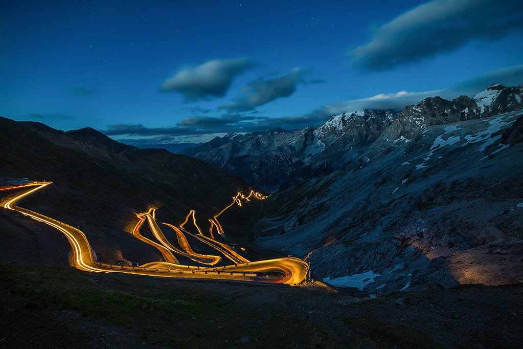 Curvy mountain road of Stelvio Pass illuminated by vehicle lights at night, surrounded by rugged alpine peaks and a deep blue twilight sky in the Italian Alps.
