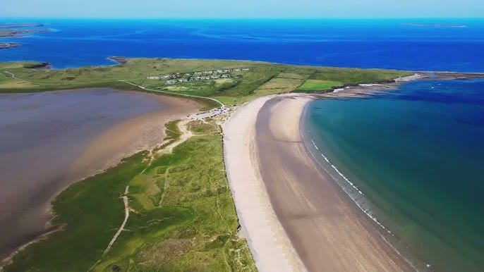 Aerial view of Streedagh Strand in Ireland, showing a long sandy beach stretching between the Atlantic Ocean and an inland lagoon, with grassy fields and a small cluster of houses nearby under a clear blue sky.