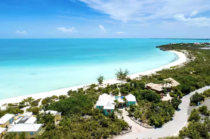 Aerial view of Taylor Bay Beach in Turks and Caicos showing turquoise shallow waters, a white sand shoreline, lush greenery, and a few beach houses with white roofs under a bright blue sky.