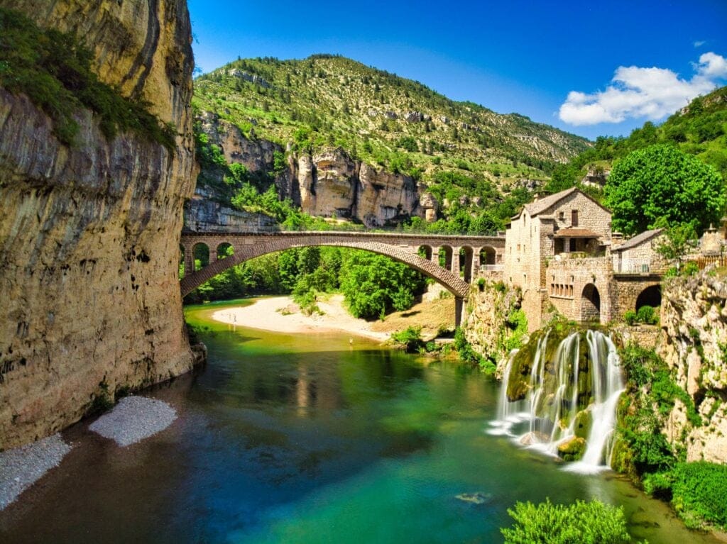 Stone bridge and waterfall beside rustic buildings in Vanoise National Park, surrounded by cliffs and forest.