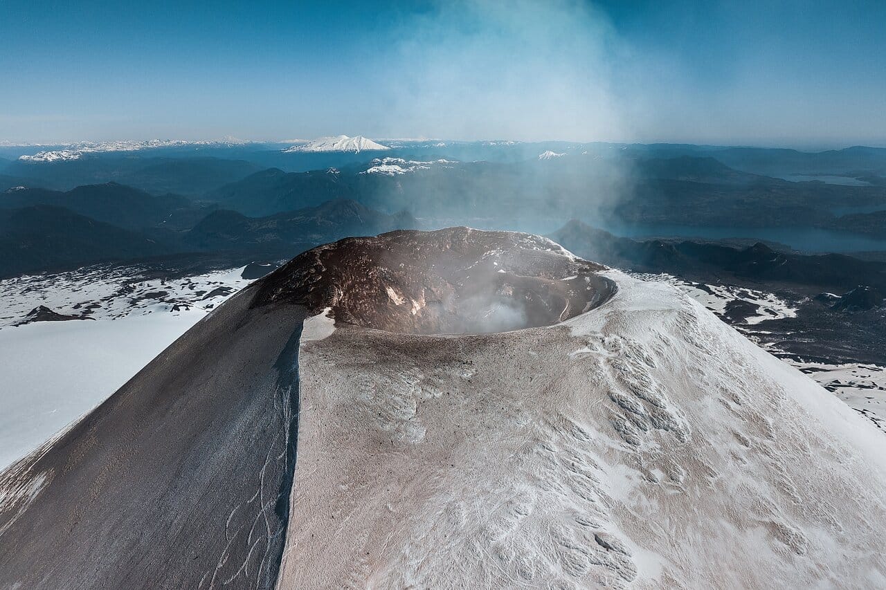 Aerial view of the smoking crater of snow-covered Villarrica Volcano in Chile, with surrounding mountains, lakes, and glaciers under a clear blue sky.
