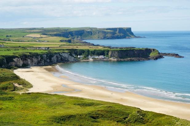 Scenic view of White Strand Beach in Miltown Malbay, Ireland, featuring a wide sandy shoreline, calm blue waters, dramatic rocky cliffs, and rolling green fields under a partly cloudy sky.