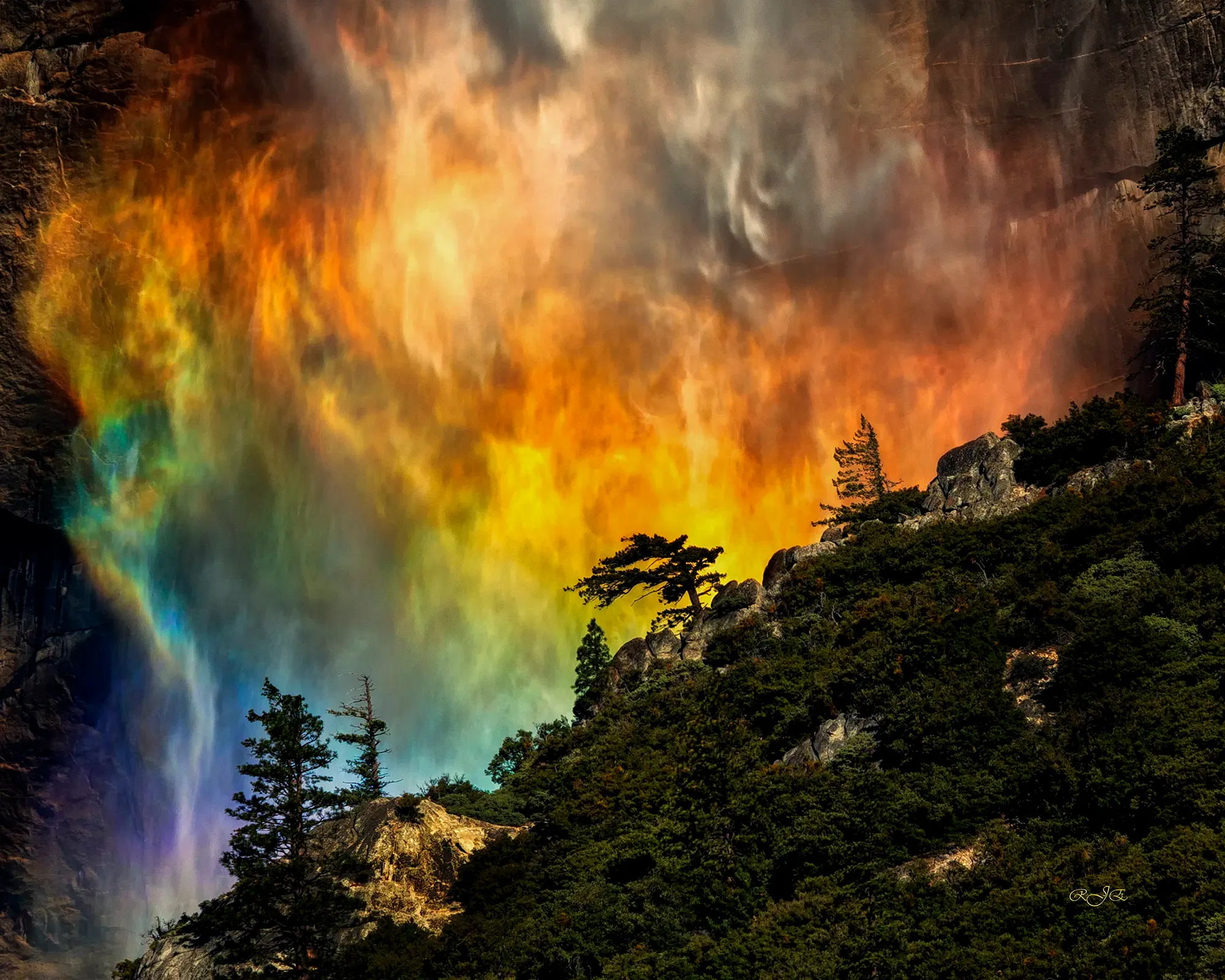A rainbow appears in the mist of a powerful waterfall against Yosemite’s rugged cliffs and pine-covered slopes.