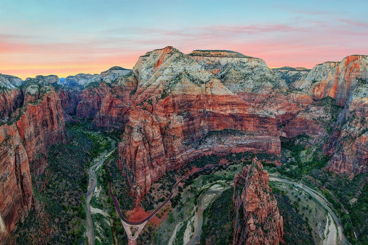 Aerial view of Zion National Park's red cliffs, winding river, and canyons with layers of sandstone glowing under a pastel sunset.
