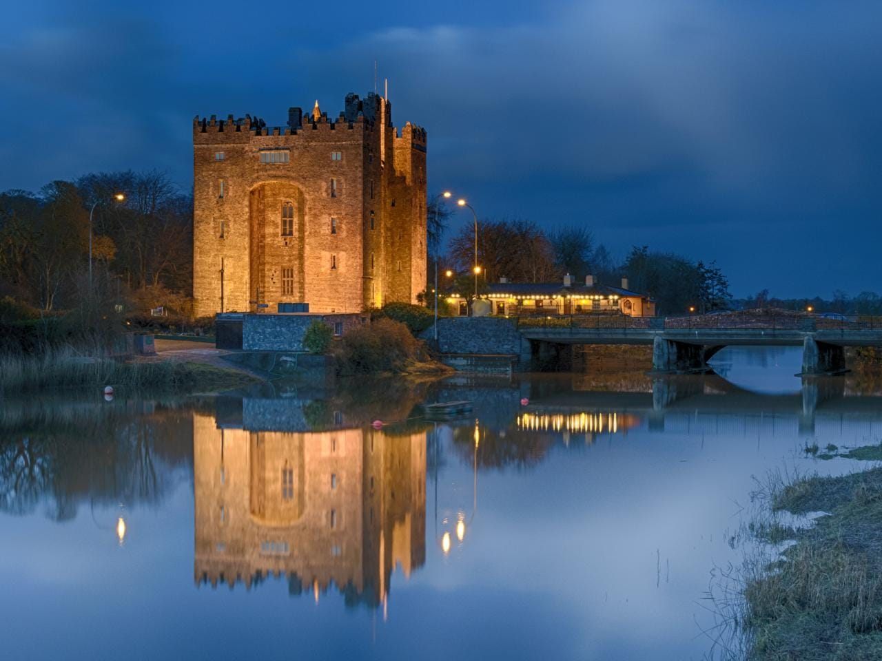 Bunratty Castle in Ireland illuminated at night, with golden lights reflecting off the calm river and a nearby bridge under a deep blue evening sky.