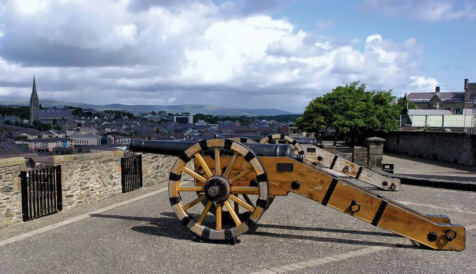Historic cannon displayed on the city walls of Derry (Londonderry), Northern Ireland, overlooking the modern cityscape with church spires and distant hills under a partly cloudy sky.