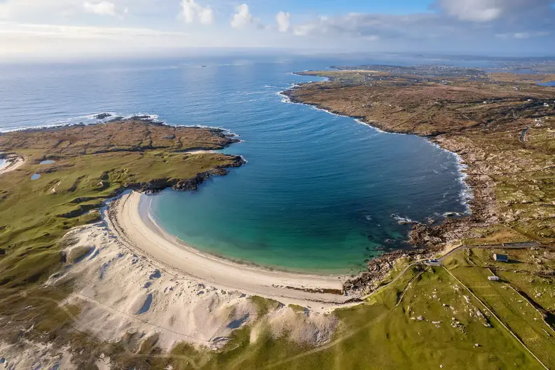 Beautiful aerial view of Dog's Bay Beach in Ireland, showcasing a curved white sandy beach bordered by bright turquoise waters and lush green hills under a partly cloudy blue sky.