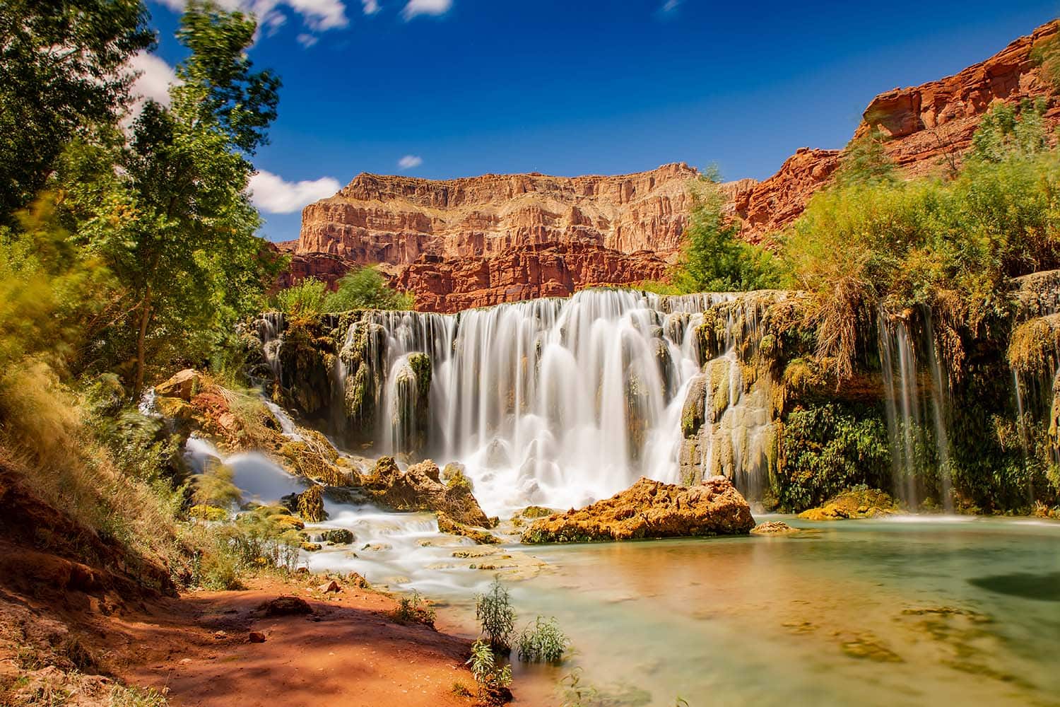 Scenic view of Beaver Falls in Arizona with tiered waterfalls, lush greenery, and red canyon cliffs under a sunny sky.and desert foliage.