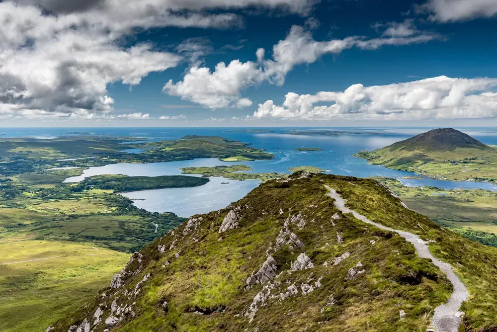 Hiking trail atop a rocky hill in Connemara National Park, Ireland, with panoramic views of lakes, green fields, and a partly cloudy sky.