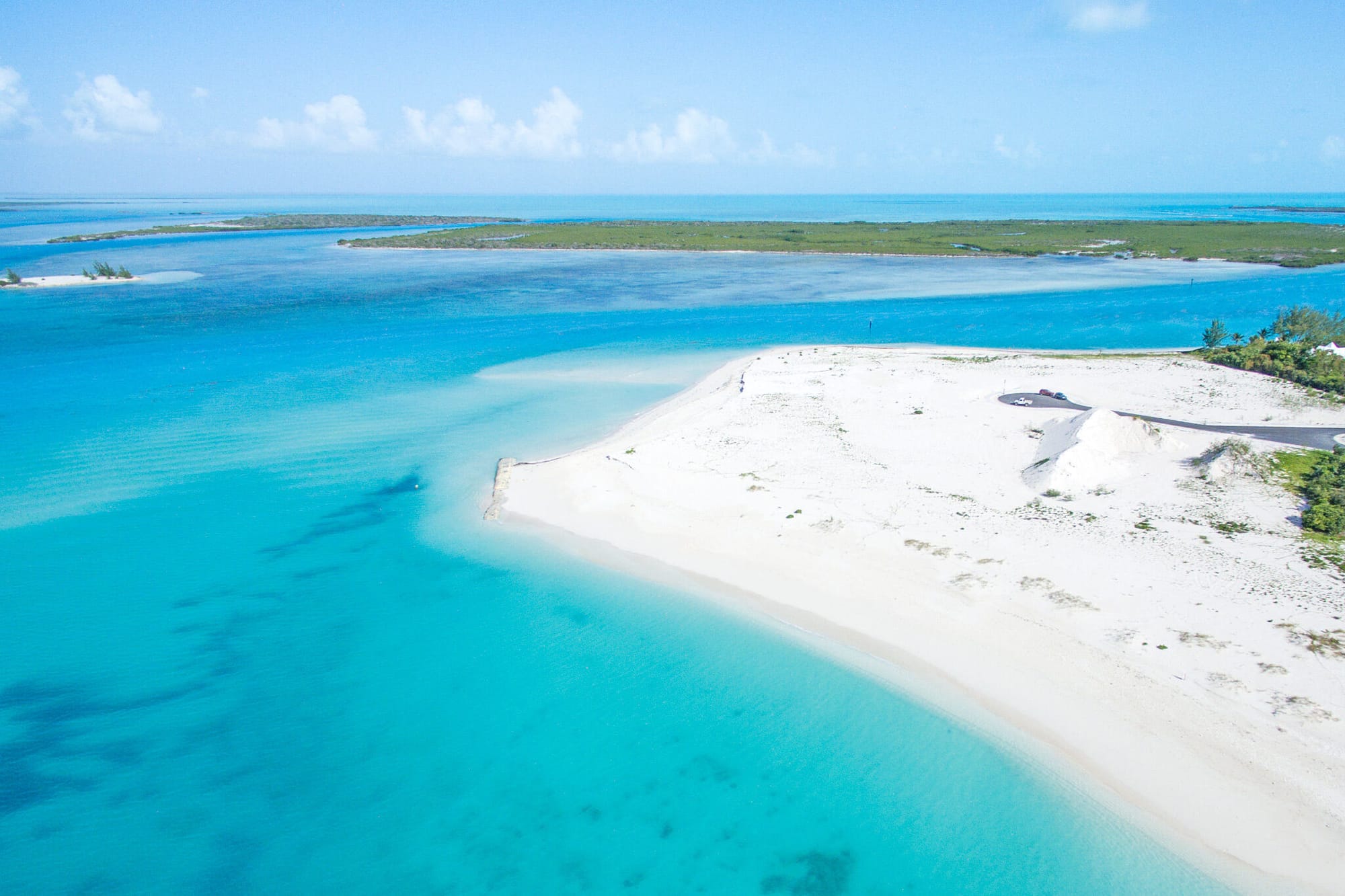 Aerial view of Leeward Beach in Turks and Caicos featuring a wide stretch of white sand, calm turquoise waters, and distant green islands under a bright blue sky with scattered clouds.