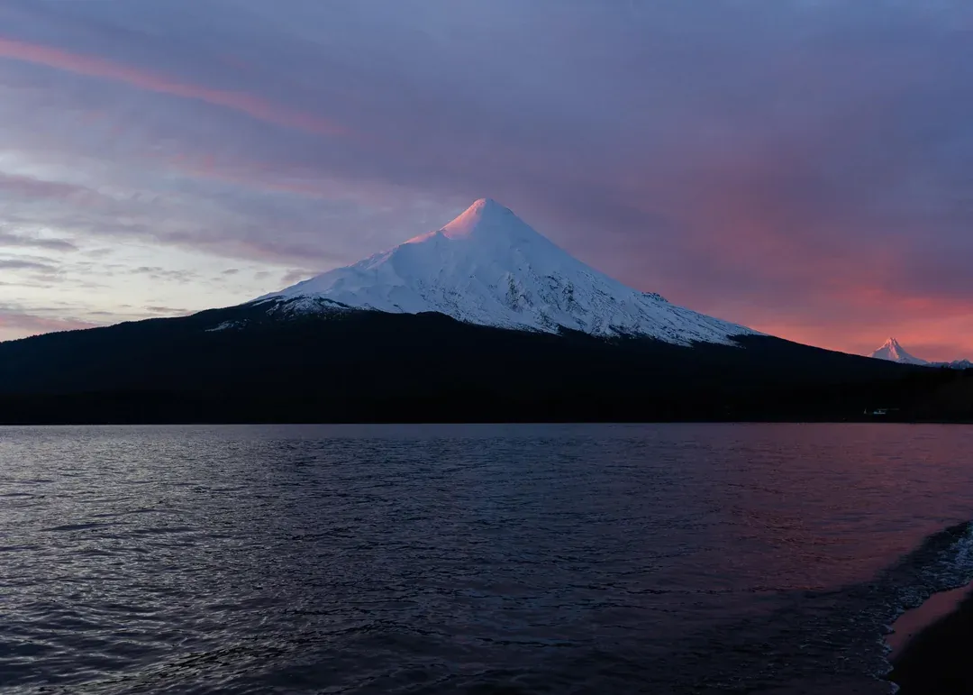 Snow-capped Osorno Volcano reflected in the calm waters of Lake Llanquihue, Chile, at sunset with a dramatic sky painted in shades of pink and purple.