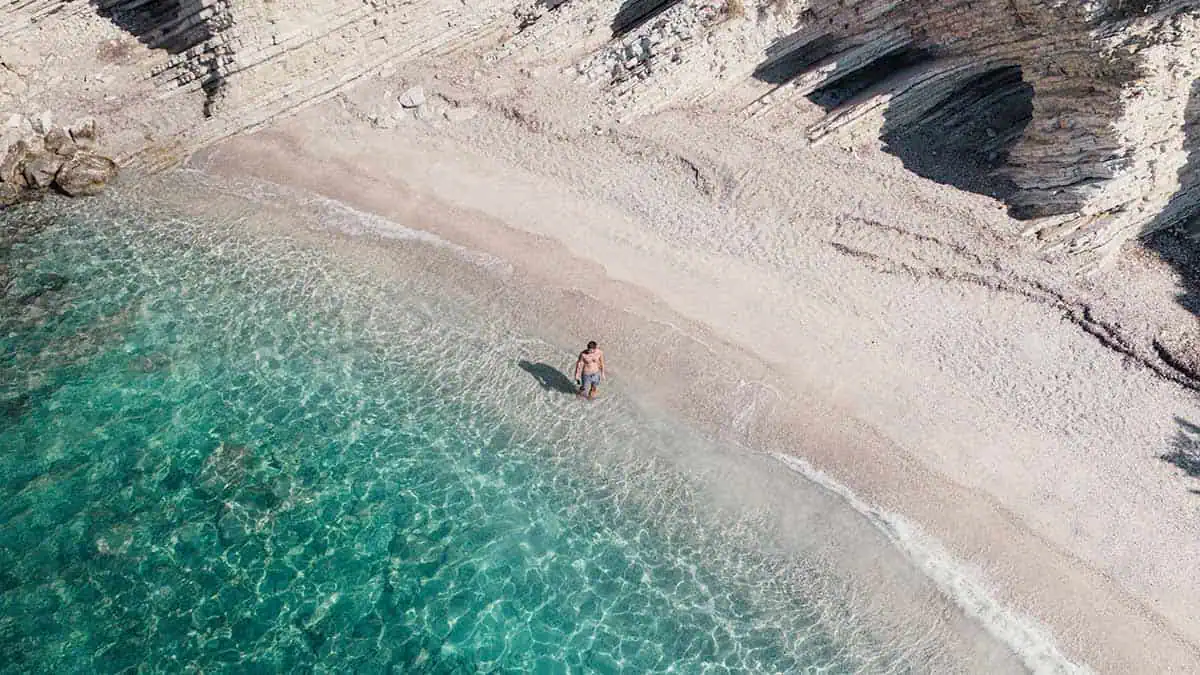 Man standing in clear turquoise water at Mirror Beach (Plazhi i Pasqyrave) in Albania, with rocky cliffs and a quiet pebbled shore.