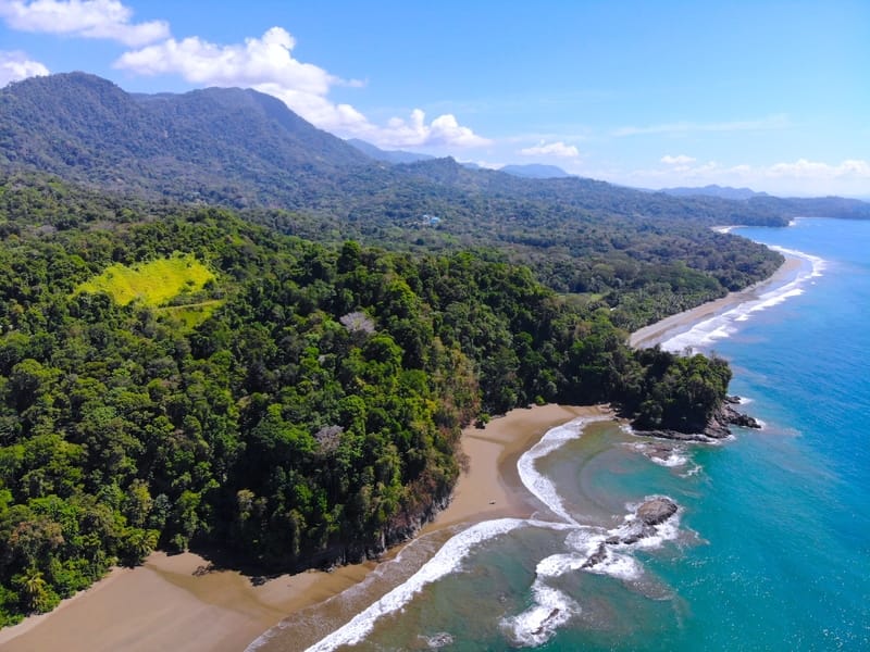 Aerial view of Playa Arco near Uvita, Costa Rica, showing a secluded sandy beach bordered by dense tropical rainforest, rocky outcrops along the shoreline, and turquoise ocean waves under a bright blue sky with scattered clouds.