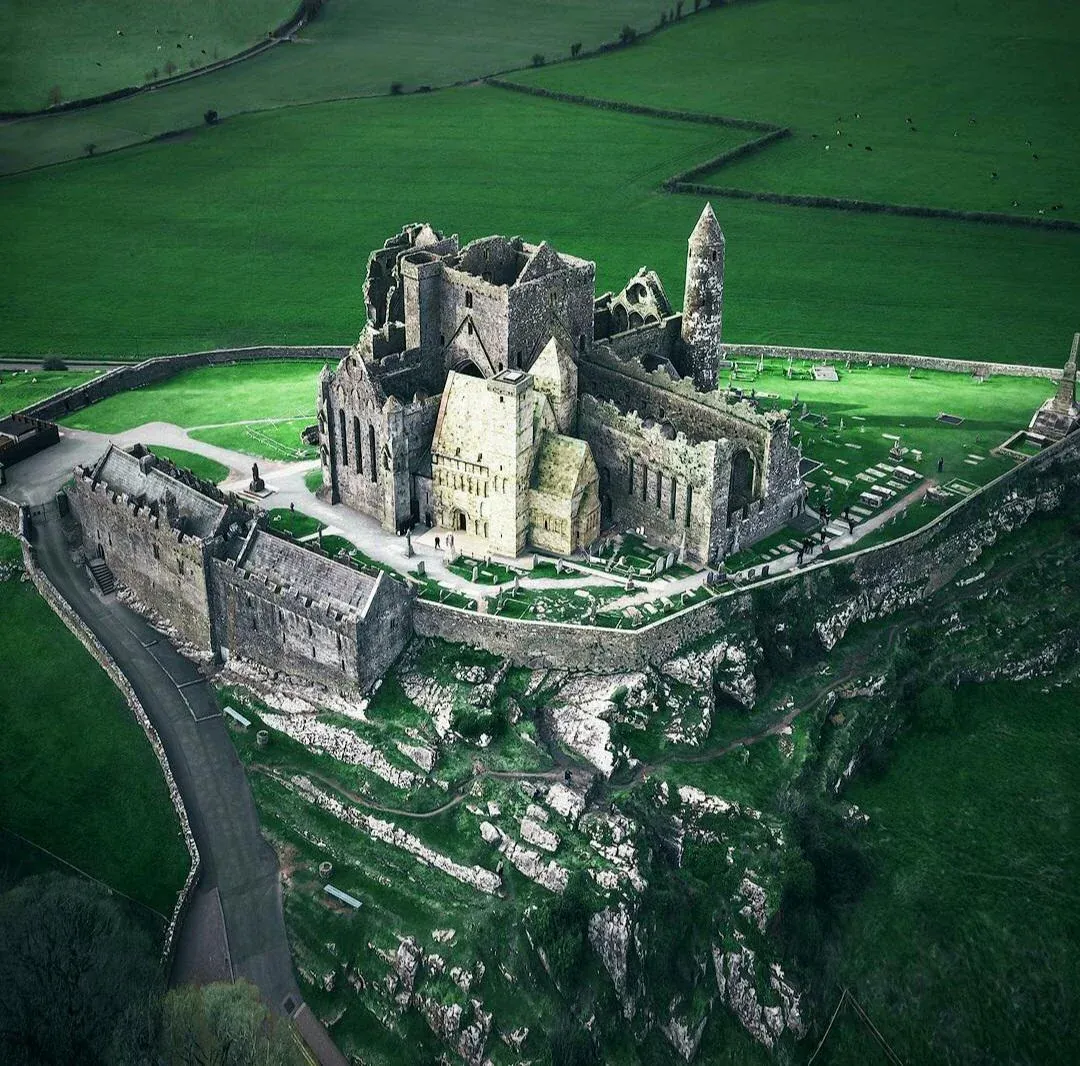 Aerial view of the Rock of Cashel in County Tipperary, Ireland, showing the ruins of medieval churches, a round tower, and stone walls perched on a rocky hill surrounded by lush green fields.