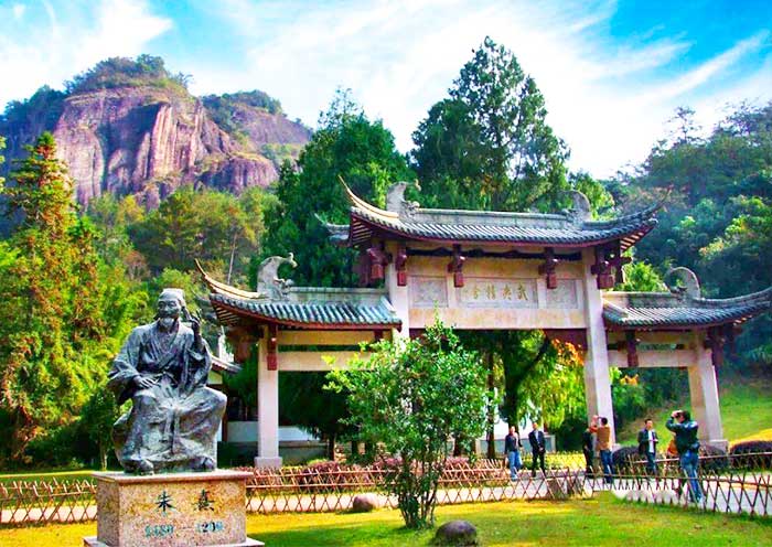 Entrance gate to Zhuxi Garden in Wuyishan, China, with a traditional Chinese archway, a statue of philosopher Zhu Xi, lush green trees, and a mountain in the background under a bright blue sky.