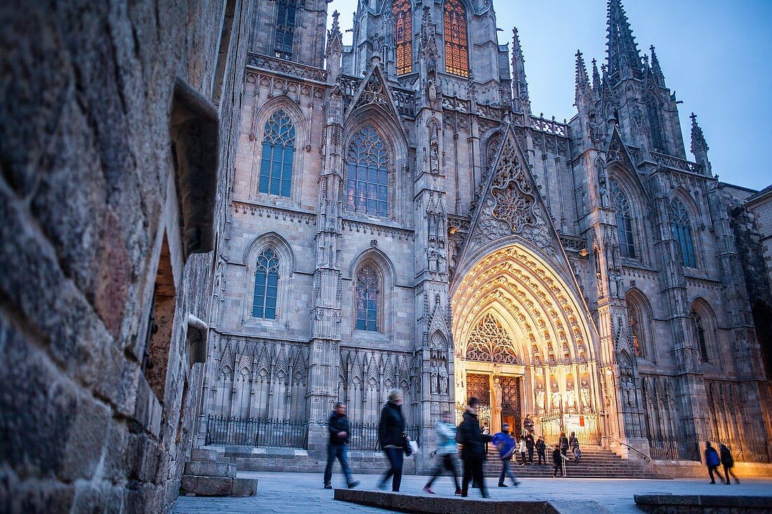 Cathedral of Holy Cross and Saint Eulalia at dusk, Gothic arches, stained glass, people walking.