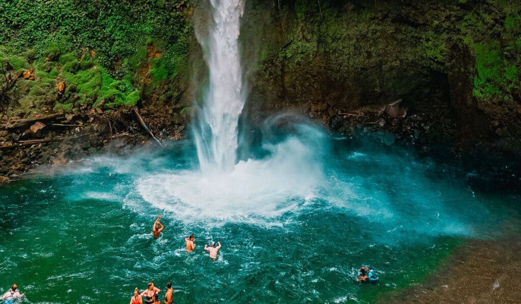 People swimming at the base of La Fortuna Waterfall in Costa Rica, where a tall stream of water crashes into a clear blue basin in a lush rainforest setting.