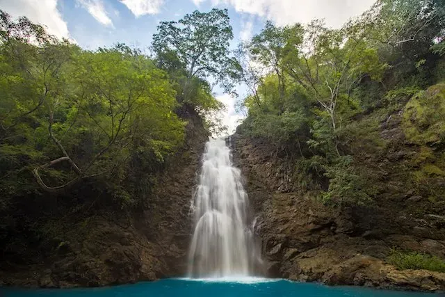 Scenic view of Montezuma Falls in Costa Rica, with water cascading down a rocky cliff surrounded by lush green tropical trees into a bright blue pool.