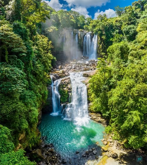 Two-tiered Nauyaca Waterfalls in Costa Rica flowing down a forested cliff into a turquoise pool, framed by dense jungle under a sunny sky.