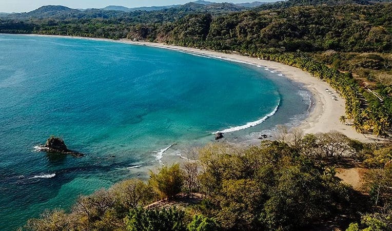 Sweeping aerial view of Playa Carrillo, Costa Rica, with a crescent-shaped bay, calm teal waters, and a quiet palm-lined beach backed by forested hills.