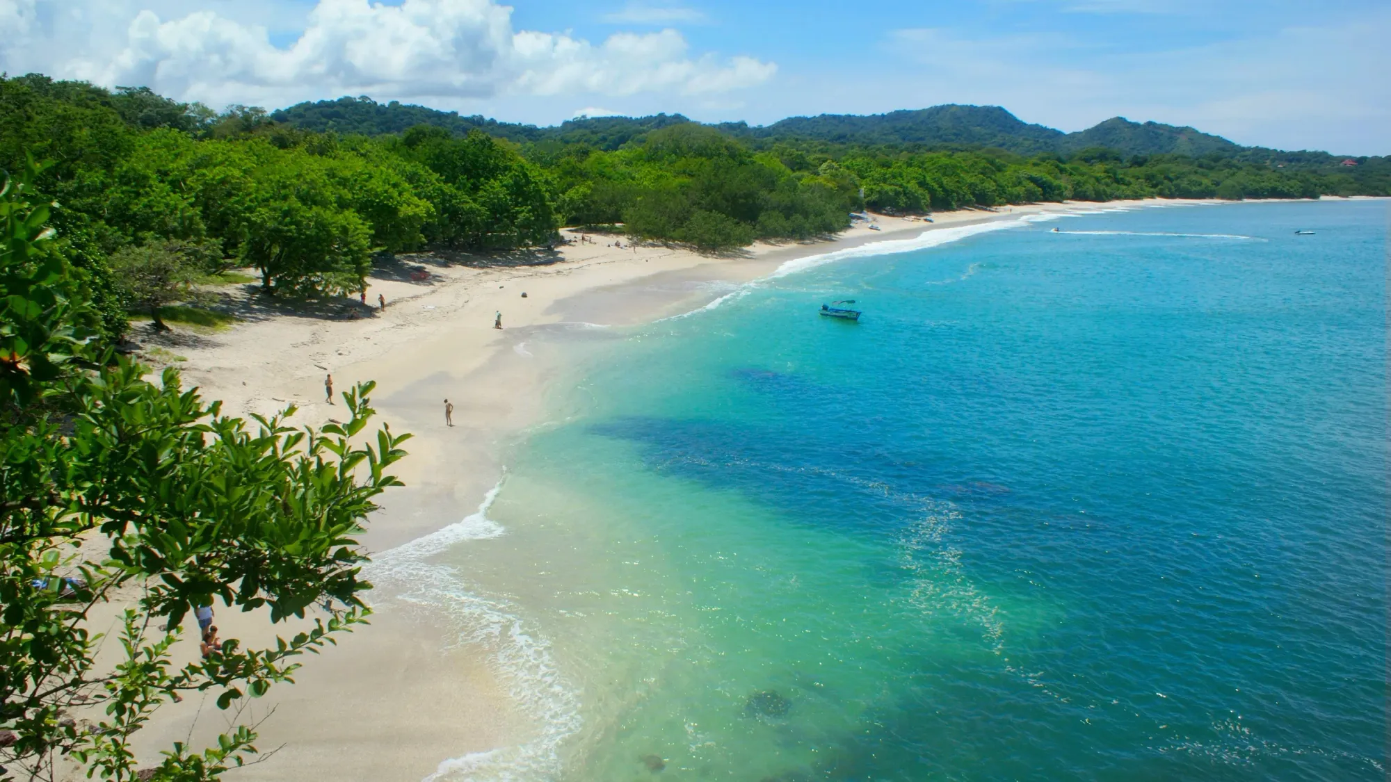 Aerial view of Playa Conchal, Costa Rica, featuring crystal-clear turquoise waters, soft white sand, and lush green forest lining the curved shoreline under a sunny blue sky.