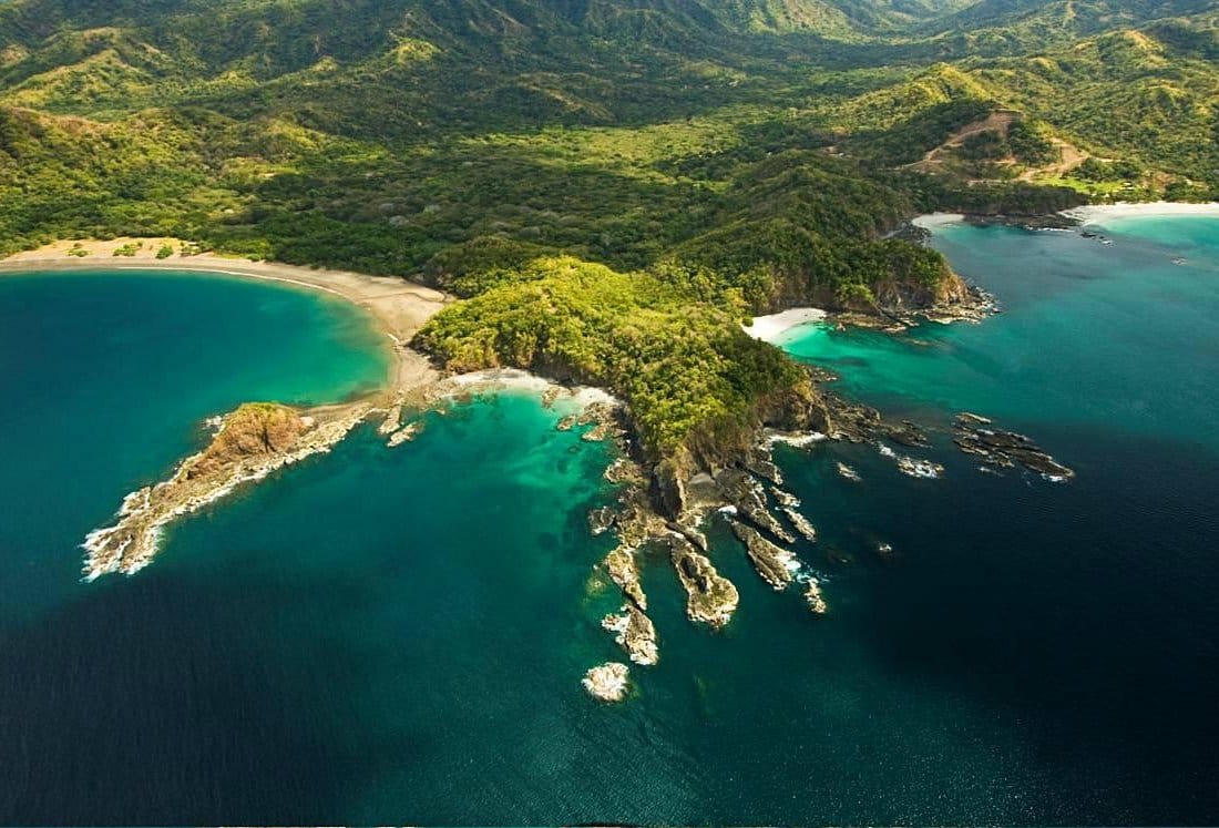 Stunning aerial view of Playa Flamingo, Costa Rica, featuring a forested peninsula between two turquoise bays with rocky outcrops and golden sandy beaches.