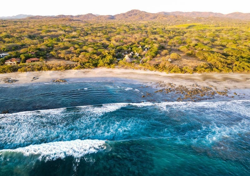 Aerial view of Playa Negra, Costa Rica, showcasing waves breaking on the dark sandy shore with forested land and rustic beach huts in the background.