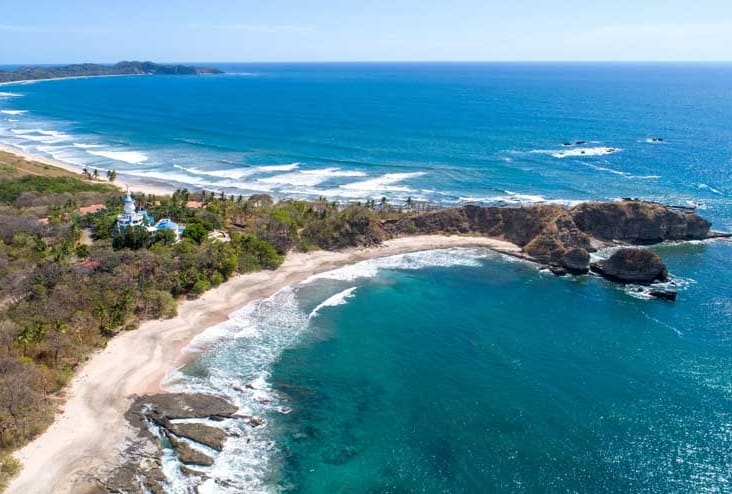 Aerial view of Playa Nosara, Costa Rica, highlighting a rugged coastline with small cliffs, rolling waves, and dry tropical forest extending inland.
