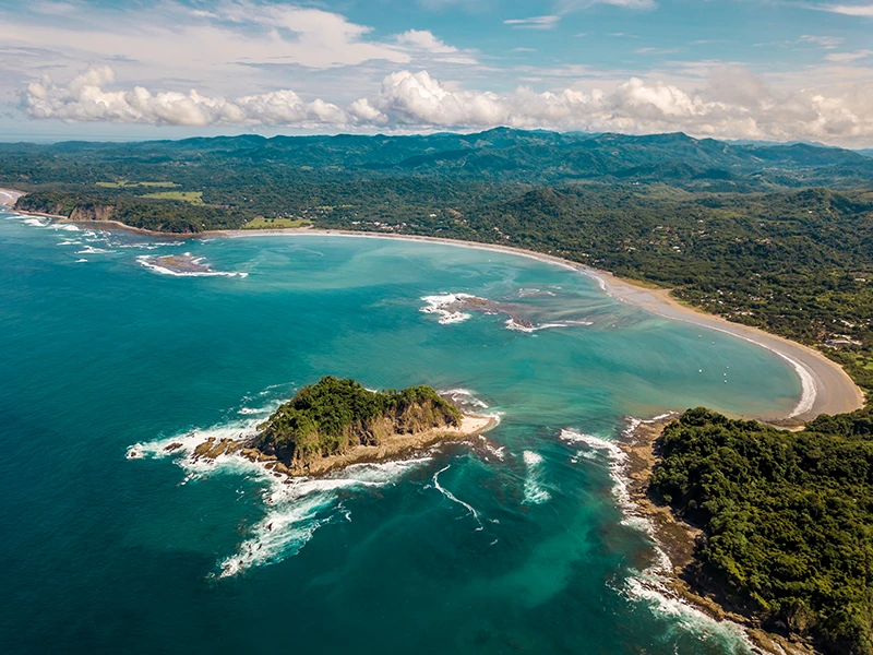 Scenic coastline of Playa Samara, Costa Rica, featuring a curved bay with shallow waves, an offshore islet, and surrounding green hills under a partly cloudy sky.