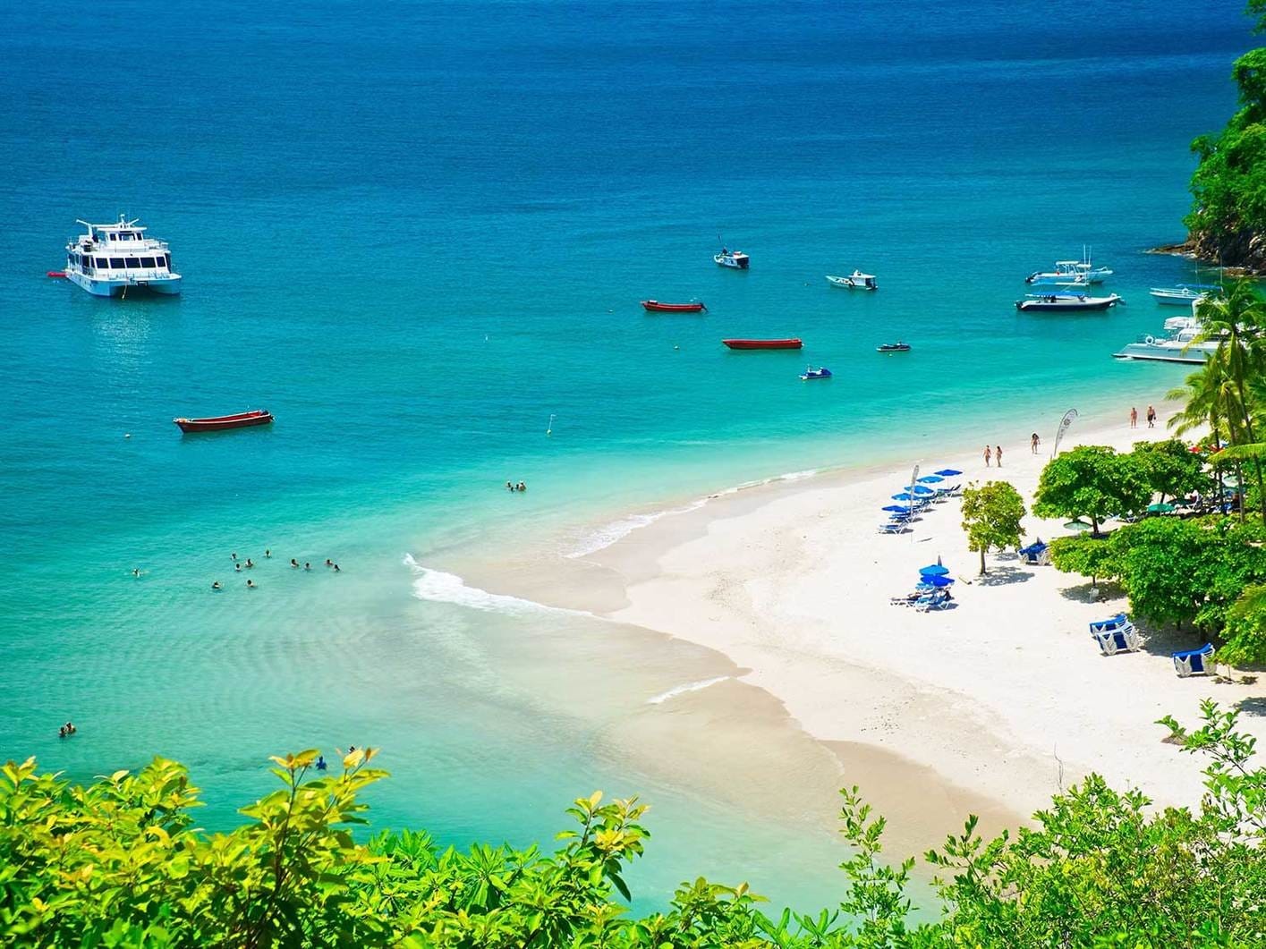 Vibrant tropical beach scene at Playa Tortuga, Costa Rica, with anchored boats and tourists swimming in turquoise waters beside white sandy shores lined with blue umbrellas and palm trees.