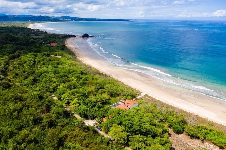 Panoramic aerial view of Tamarindo, Costa Rica, showing a wide sandy beach with gentle surf, surrounded by green hills and scattered coastal buildings.