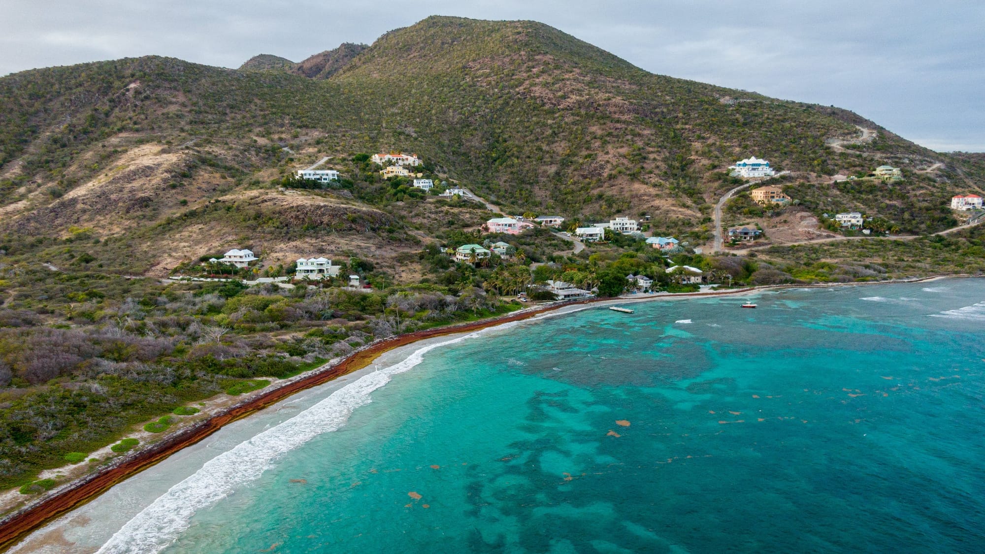   Very clear blue waters and a breach sitting in front of a good size mountain with houses on the mountain side at Turtle Beach in St. Kitts.