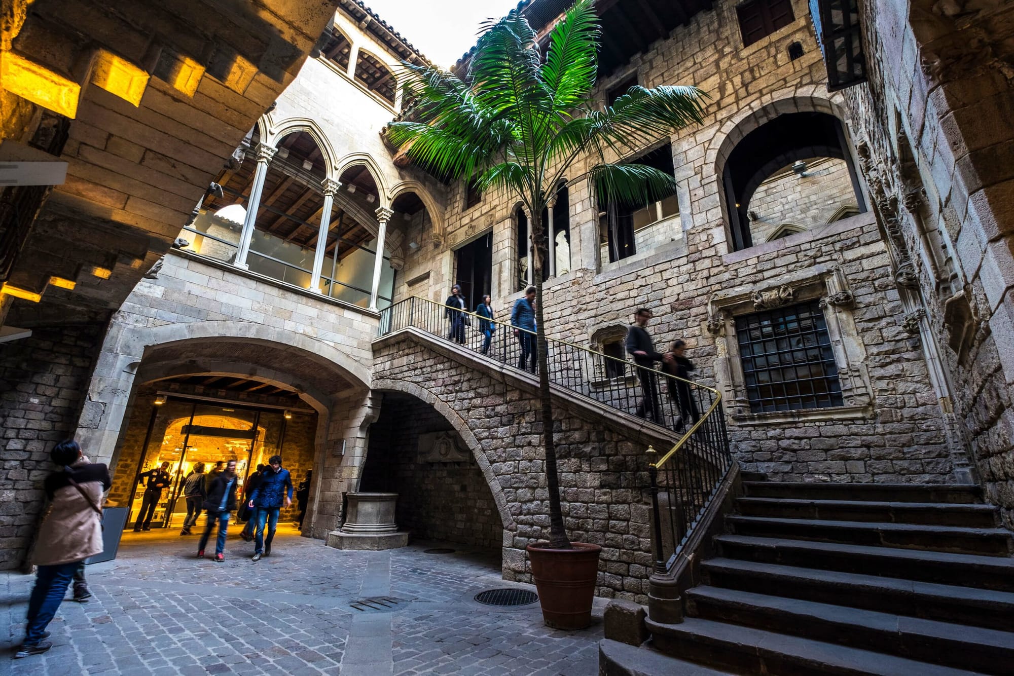 Stone arches and a palm tree with people walking in the courtyard of the Museu Picasso.
