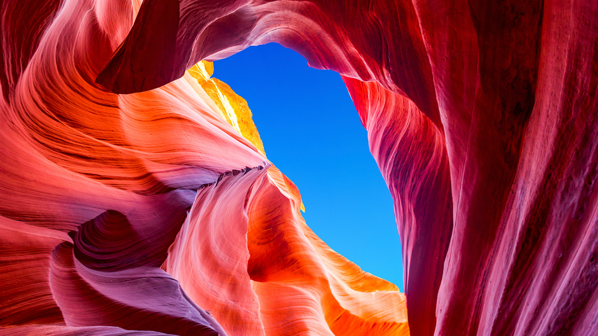 View looking up through the winding, smooth, red and orange sandstone walls of Antelope Canyon, with sunlight illuminating the curves and a vivid blue sky visible through an opening above.