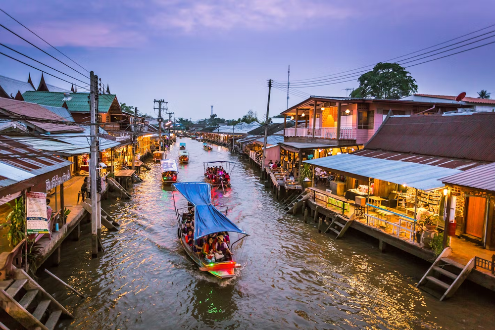 Evening view of longtail boats navigating a narrow canal lined with traditional wooden houses and local shops at the Artist’s House (Baan Silapin) area in Bangkok, Thailand, under a purple sunset sky.