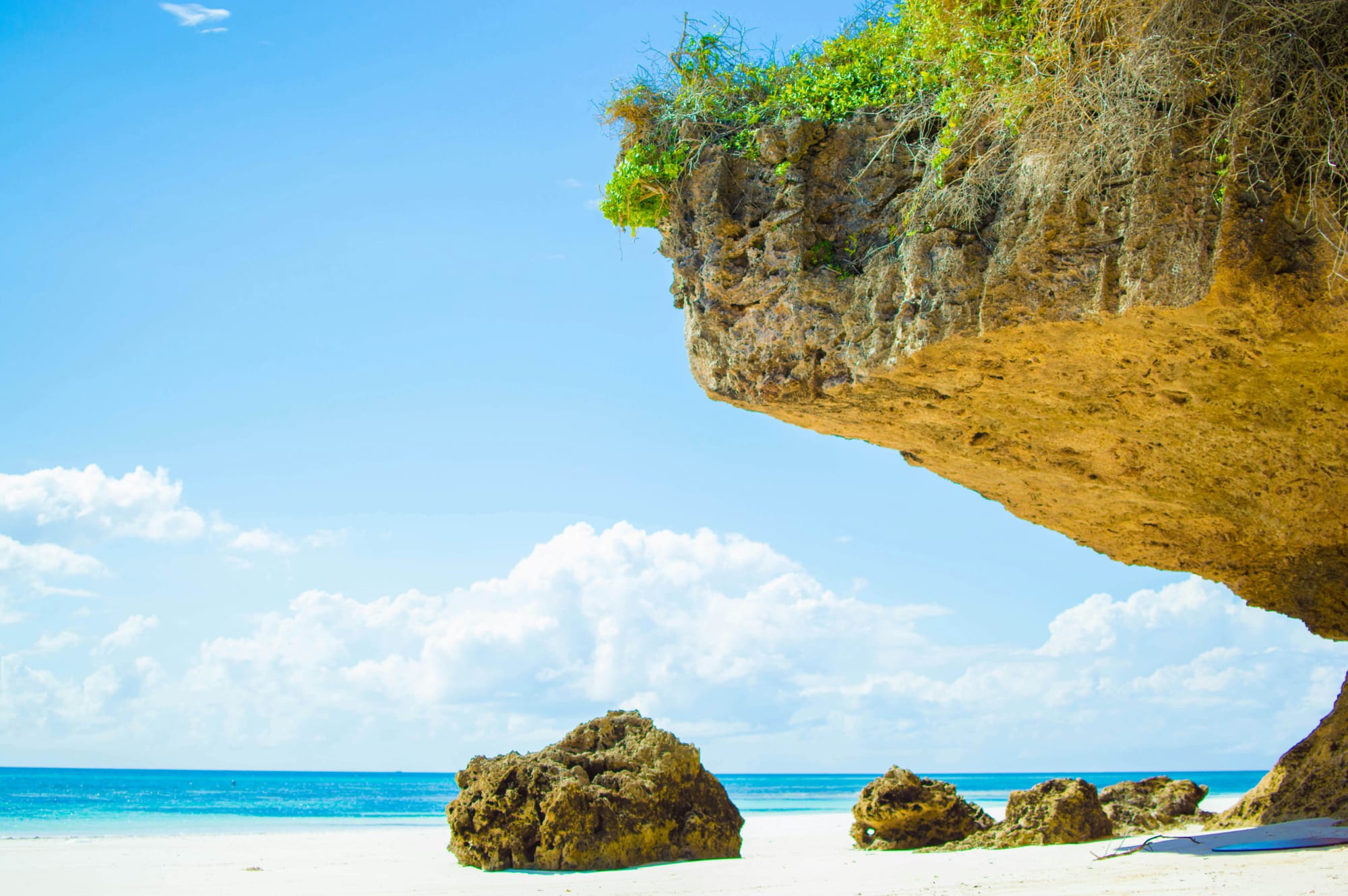Close-up view of a rocky overhang on Bamburi Beach, Kenya, with white sand, scattered rocks, and the calm, blue Indian Ocean in the background under a clear sky.