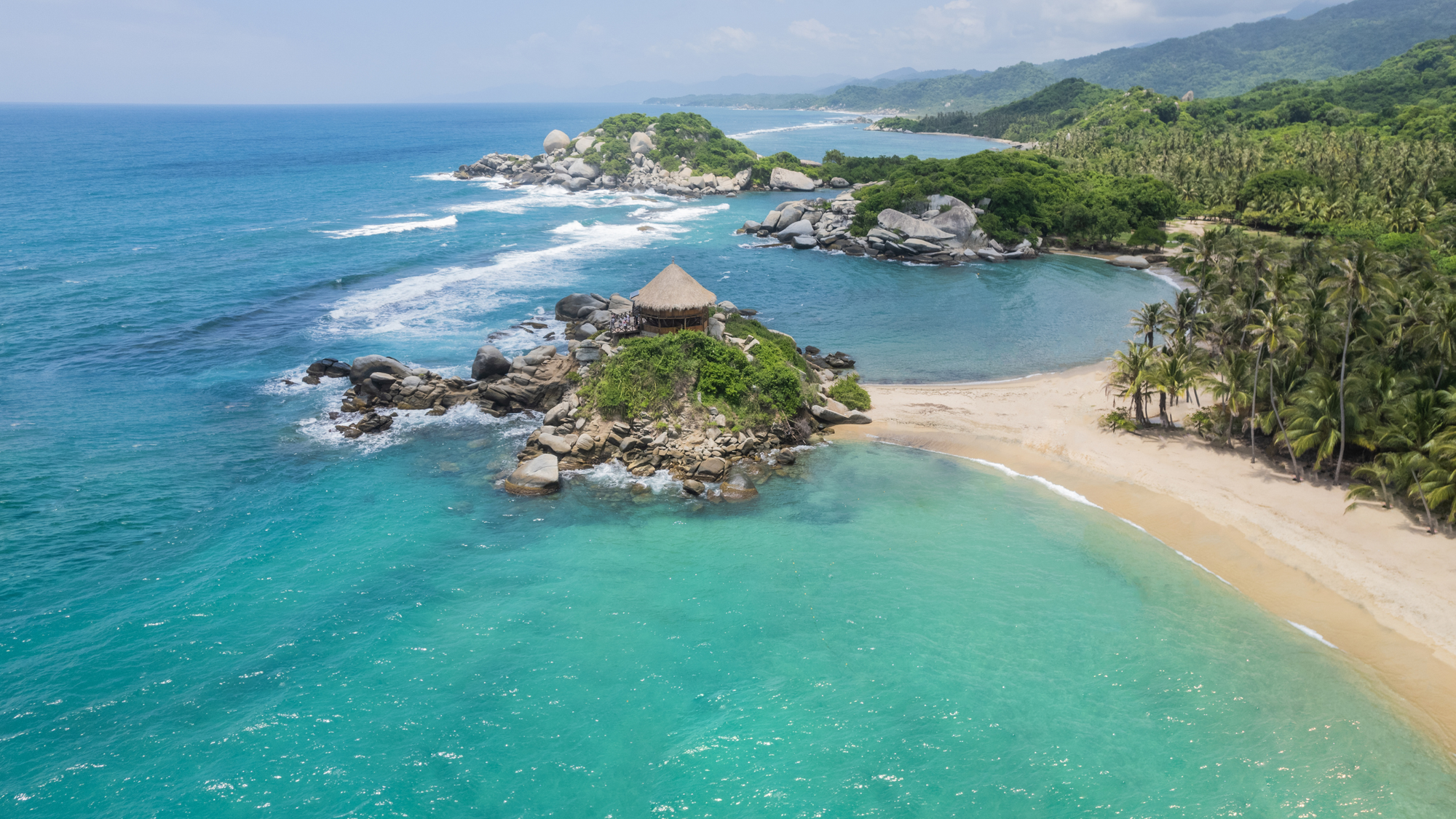 Thatched-roof hut on a rocky outcrop surrounded by turquoise waters and palm-lined beaches at Tayrona National Park on Colombia’s Caribbean coast.