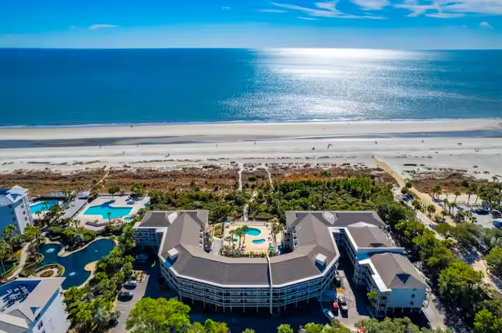 Aerial view of Coligny Beach on Hilton Head Island, South Carolina, featuring a large oceanfront resort with pools, landscaped grounds, and a wide sandy beach facing the Atlantic Ocean under a bright blue sky.