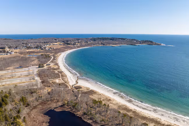 View of Crescent Beach in Cape Elizabeth, Maine, showing waves lapping against a curved sandy shoreline with one person walking along the beach near rocky outcrops.
