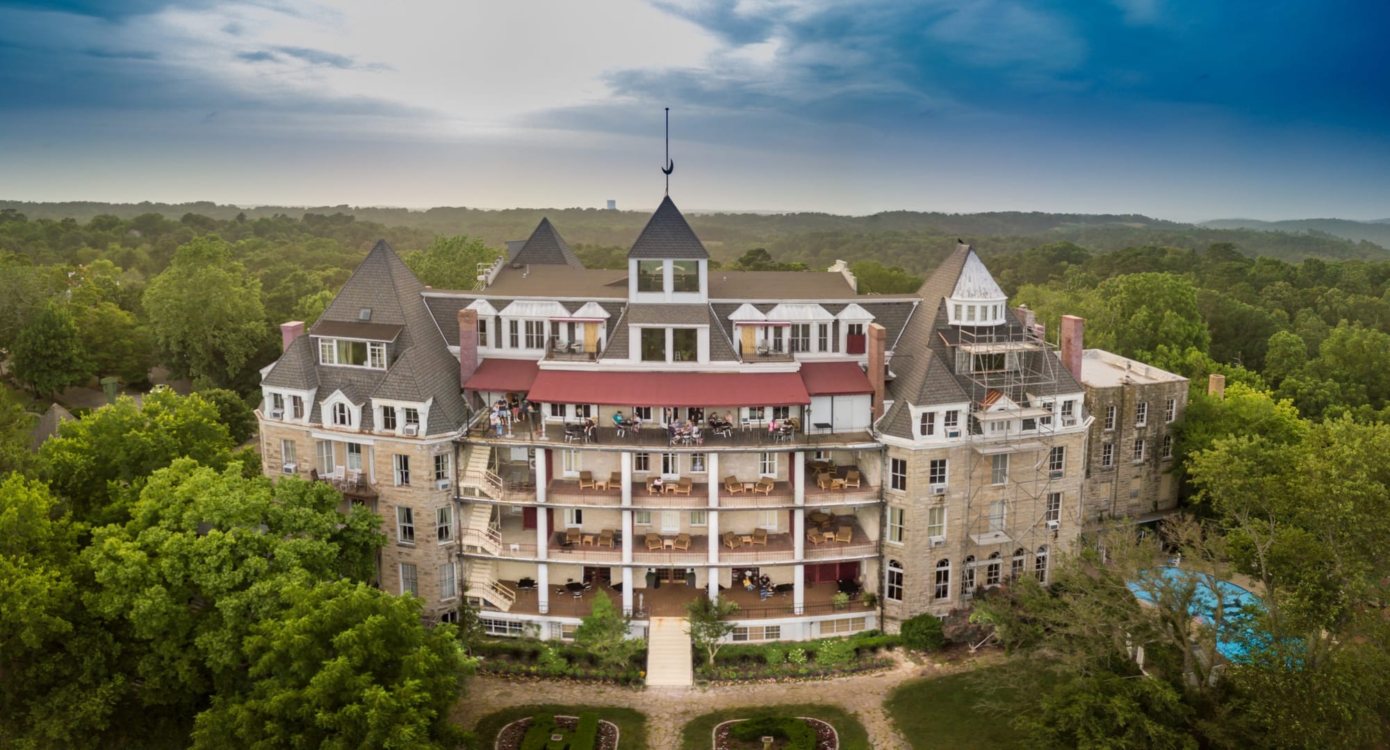 Aerial view of the historic Crescent Hotel in Eureka Springs, Arkansas, surrounded by lush green forest under a partly cloudy sky.