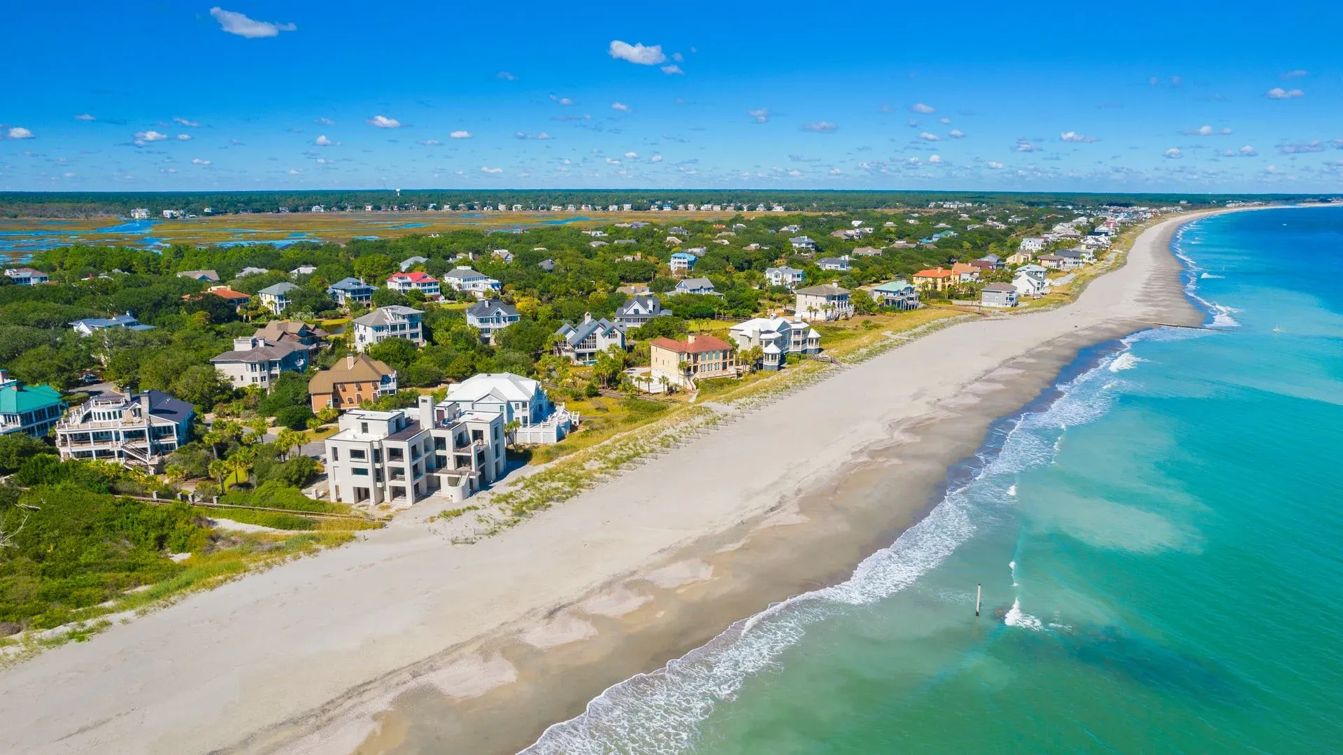 Aerial view of Debordieu Beach near Georgetown, South Carolina, showing oceanfront homes, a wide sandy shoreline, and clear turquoise waters under a bright blue sky.