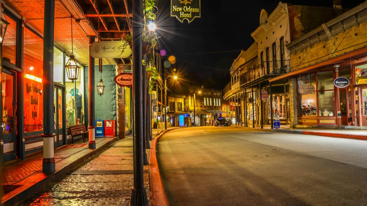 Night view of historic downtown Eureka Springs, Arkansas, featuring lit storefronts, vintage signs, and a quiet, curved street.