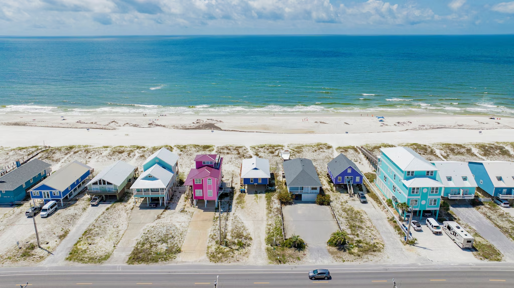 Colorful beachfront houses lined up along Drift Inn Beach in Maine, facing a wide sandy shoreline and calm blue ocean under a partly cloudy sky.