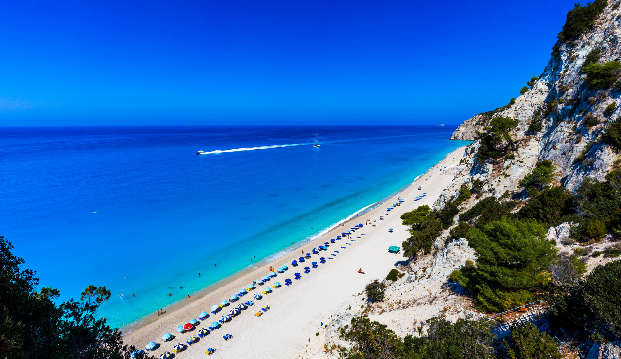 A long stretch of Egremni Beach on Lefkada Island, Greece, lined with sun umbrellas and backed by steep white cliffs, overlooking calm azure waters.
