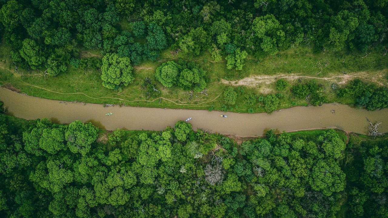 Aerial top-down view of a muddy river winding through the dense, green forest of El Impenetrable National Park in Argentina with small kayaks on the water.