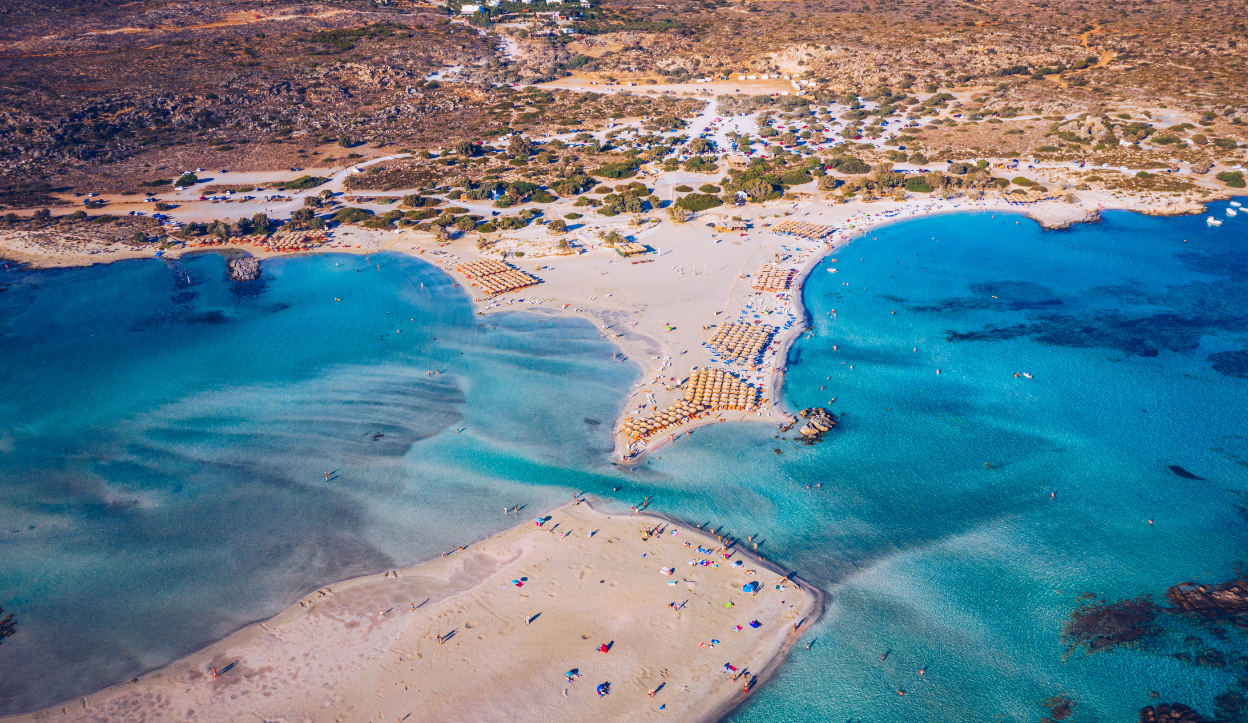 Aerial photo of Elafonissi Beach in Crete, Greece, with shallow turquoise lagoons, soft pinkish sand, and a narrow sandbar connecting to a small offshore islet.  Let me know if you’d like these optimized for SEO with specific keywords or fo