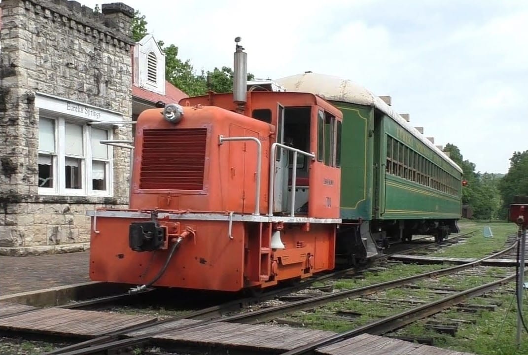 Vintage red and green train at the Eureka Springs & North Arkansas Railway station, with stone buildings and trees in the background.