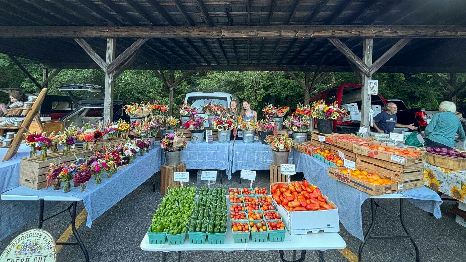 Colorful display of fresh produce and flower bouquets at the Eureka Springs Farmers Market, with vendors and customers under a wooden pavilion.