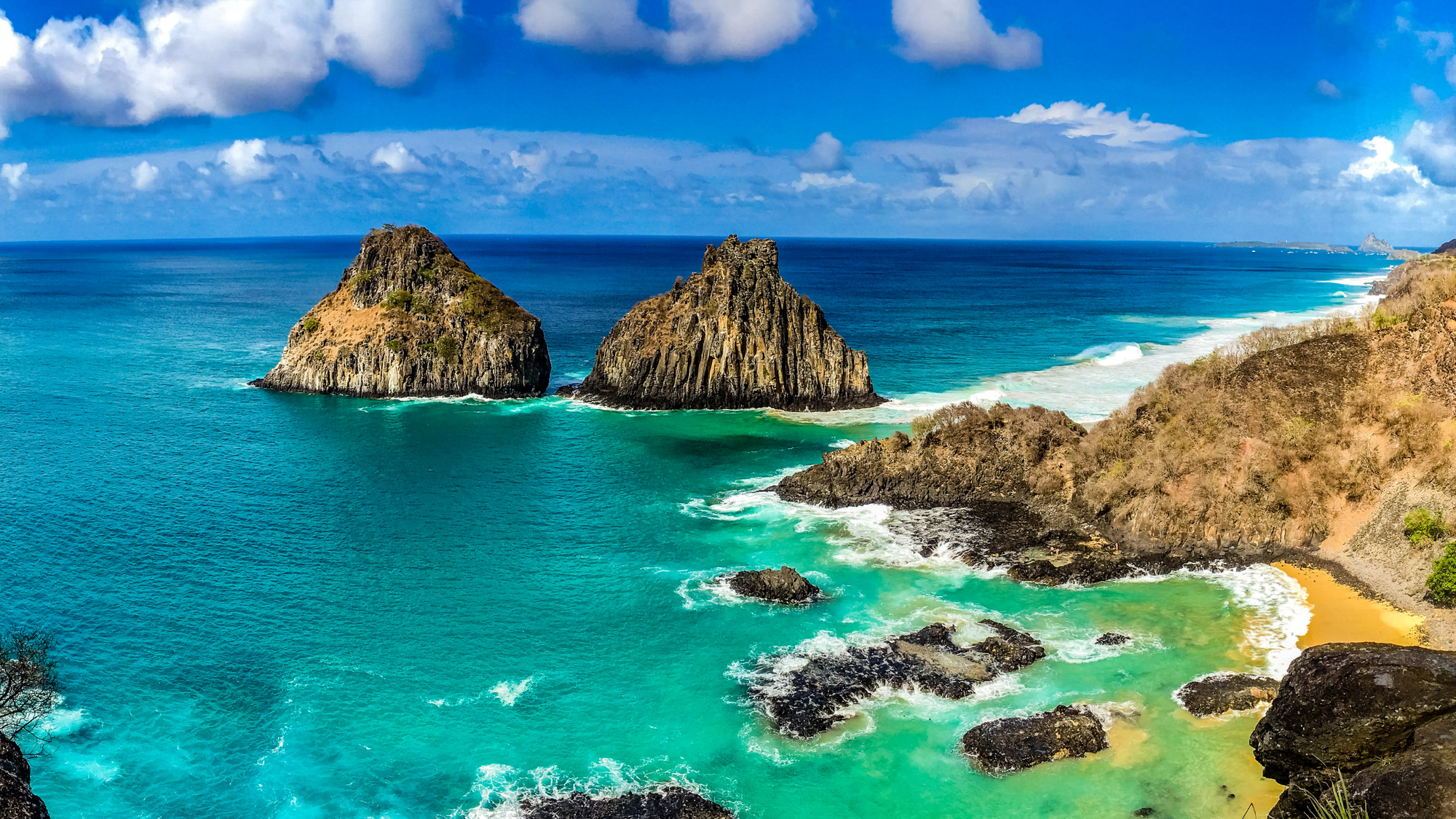 Stunning coastal view of Fernando de Noronha Marine National Park in Brazil, featuring turquoise waters, dramatic rocky islets, and golden sandy beaches under a bright blue sky with scattered clouds.